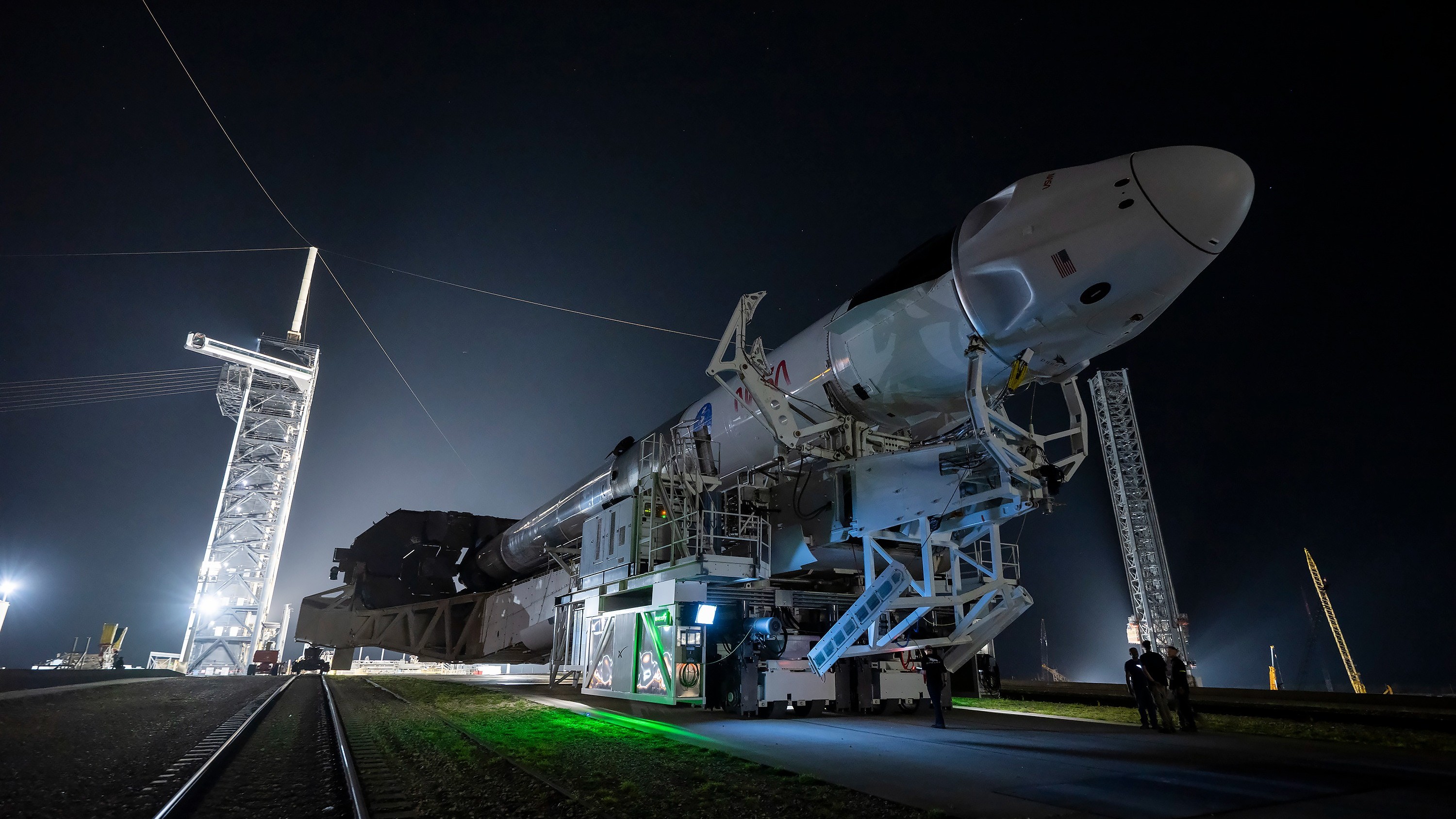 A tall white rocket is rolled on its back toward a launch tower, light with bright lights in a dark hazy night.