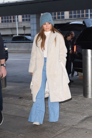 J.Lo wears a blue beanie, ivory shearling coat, white turtleneck, flare jeans, and white boots.