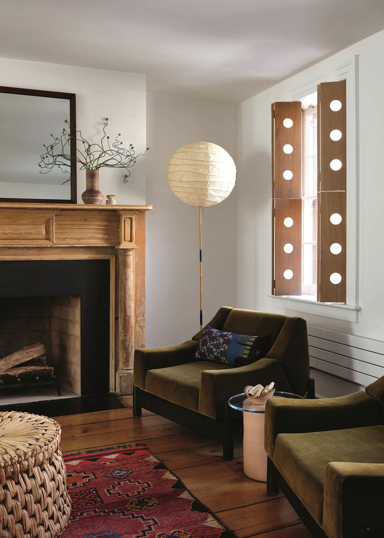 living room with timber fireplace, paper lantern on floor lamp, green armchairs with red rug, woven coffee table, and timber shutters with holes in them
