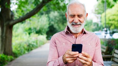 An older man smiles as he looks at his phone while walking in a park.