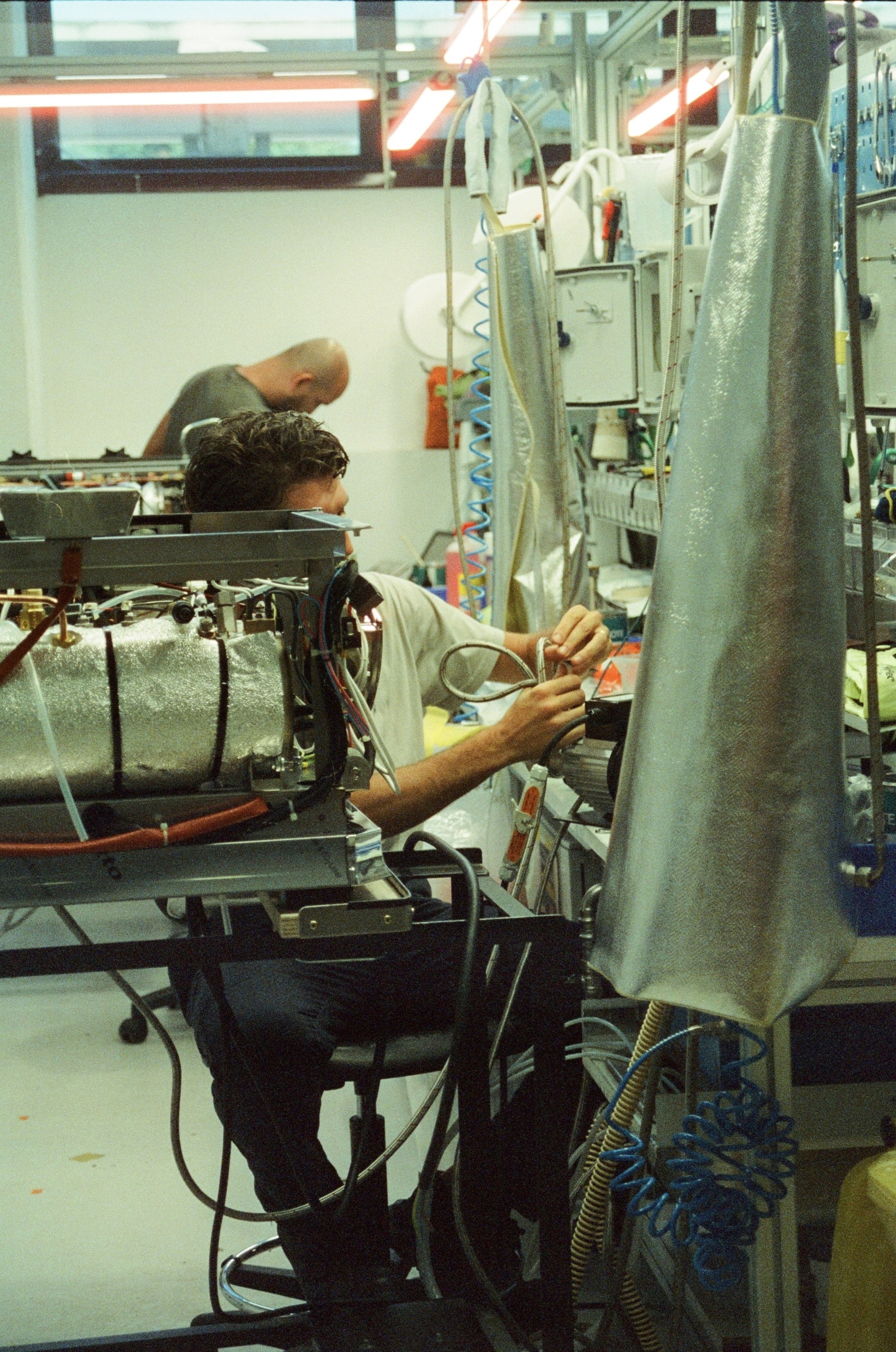 Image of a man in a white shirt and black pants sitting on a desk chair in a white factory, putting together a coffee machine.
