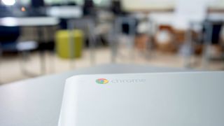 A photo of a silver Google Chromebook on a desk, with a blurred classroom in the background