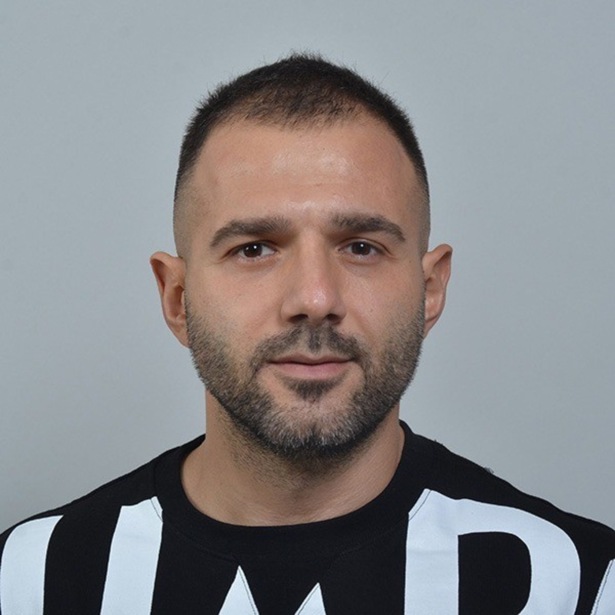 A picture of Seymen Usta, wearing a white and black shirt and standing in front of a gray background