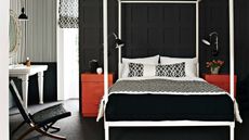 A black bedroom with wall panelling, a large white four poster bed, and two red chest of drawers to either side, with matching black lamps. To the left is a black and white striped wall, white sink, large window, circular mirror, and black chair.