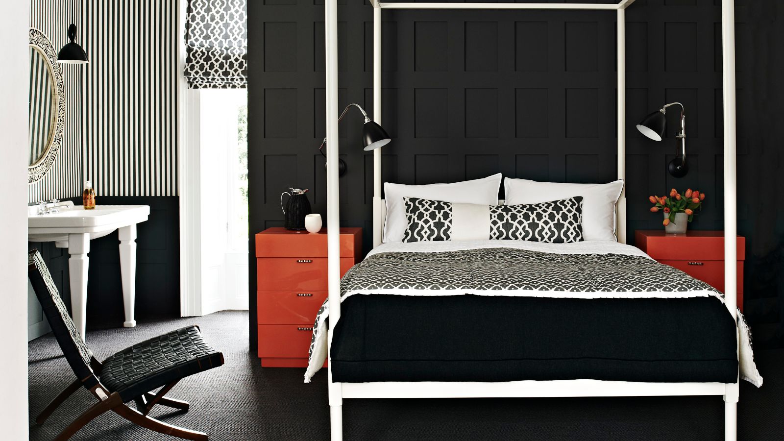 A black bedroom with wall panelling, a large white four poster bed, and two red chest of drawers to either side, with matching black lamps. To the left is a black and white striped wall, white sink, large window, circular mirror, and black chair.