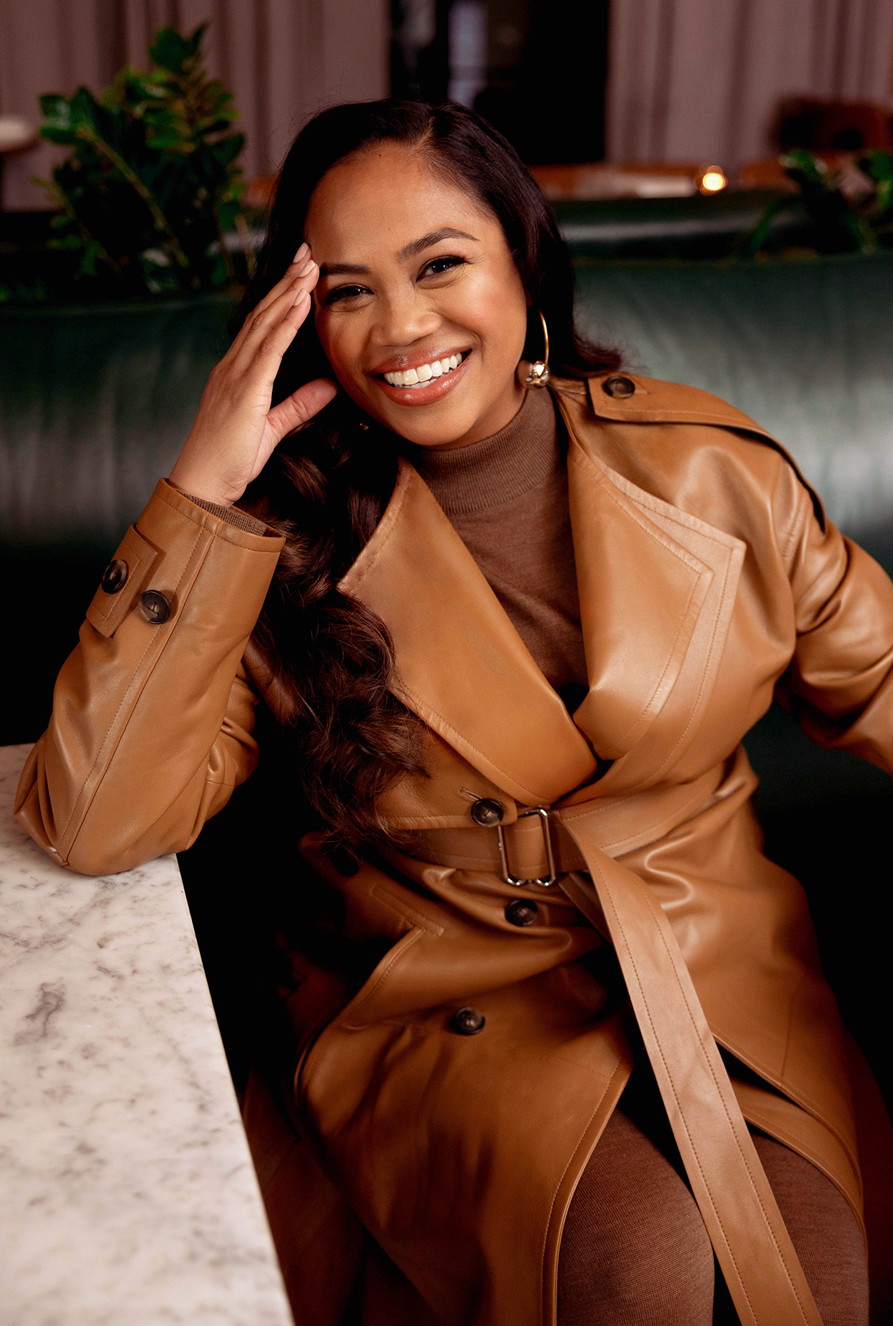 Zinzi Coogler laughing with her elbow on a marble table, wearing a camel leather jacket