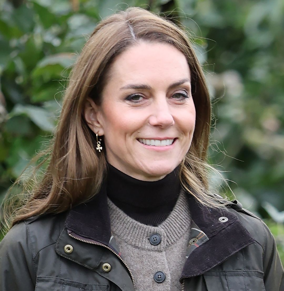 Kate Middleton wearing a green jacket and brown cardigan in front of trees
