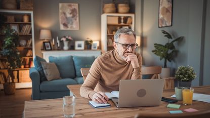 An older man looks thoughtful as he looks at his laptop while sitting at his dining room table.