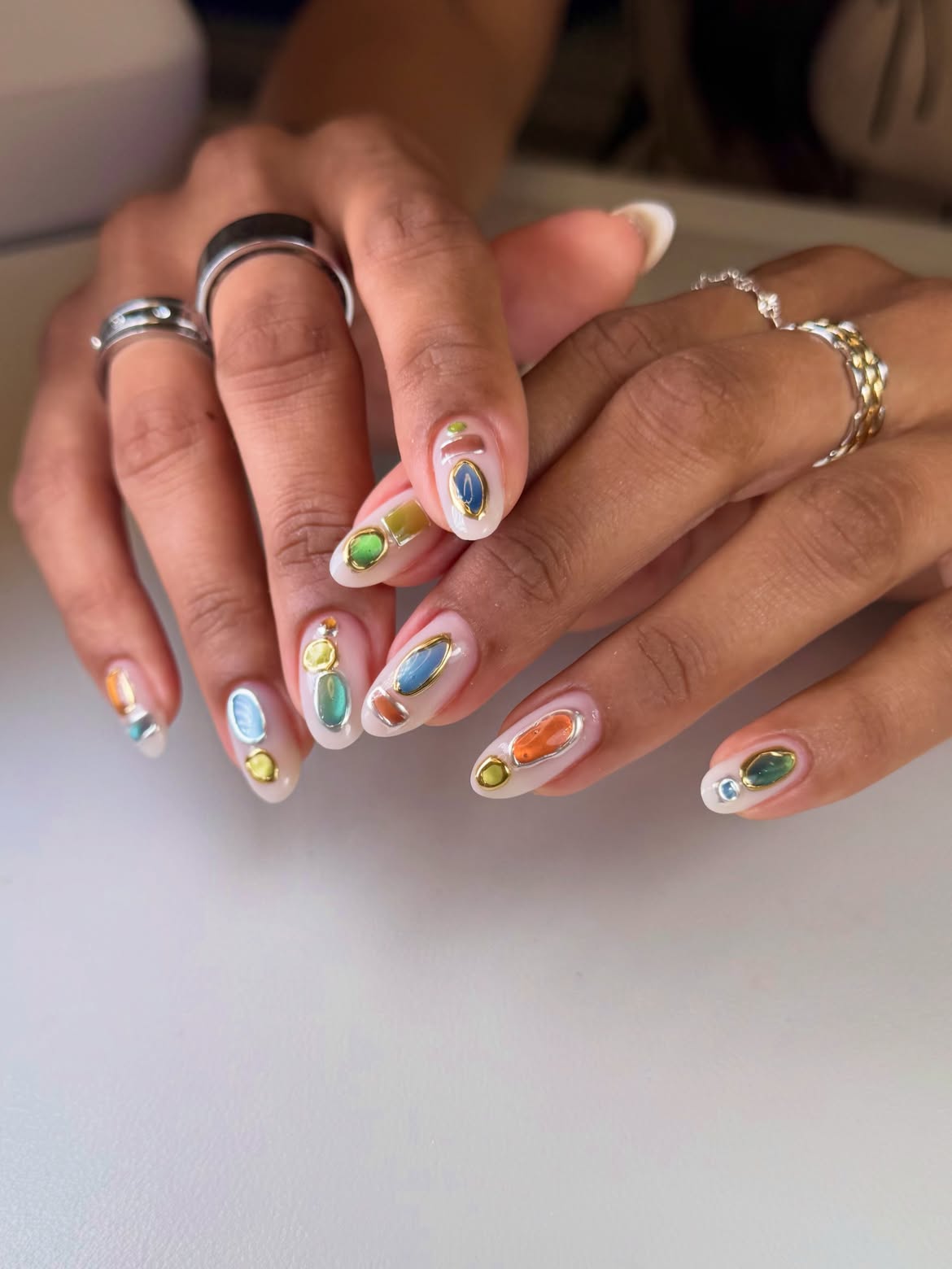 Milky almond nails decorated with translucent jewel-toned shapes outlined in raised metallic gold