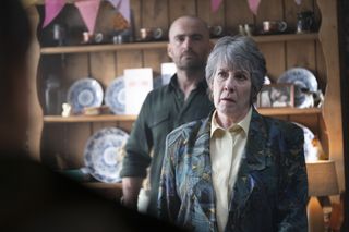 Grace Bain (Phyllis Logan) stands inside her kitchen, in front of a cabinet filled with plates, birthday cards and bunting. Her son Bobby (Russ Bain) stands behind her, leaning against the cabinet.