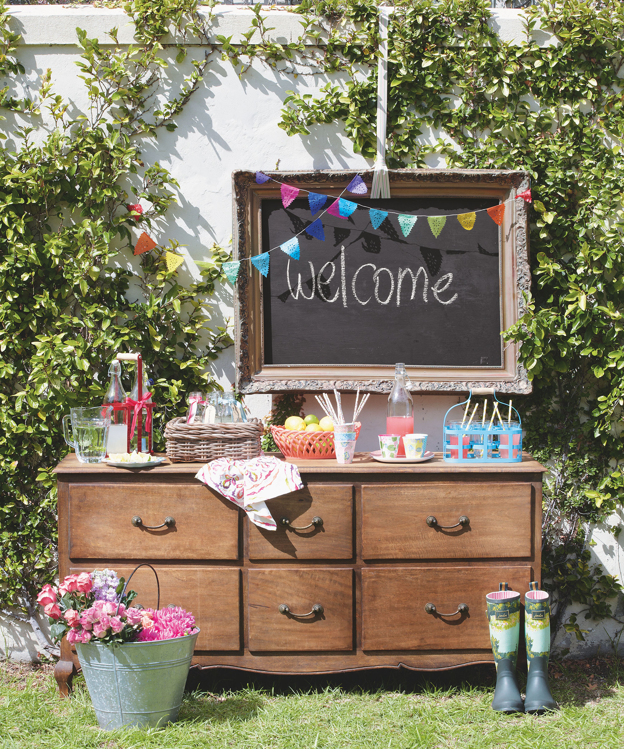 A wooden chest of drawers with food and drink on it in front of a Welcome sign in a garden