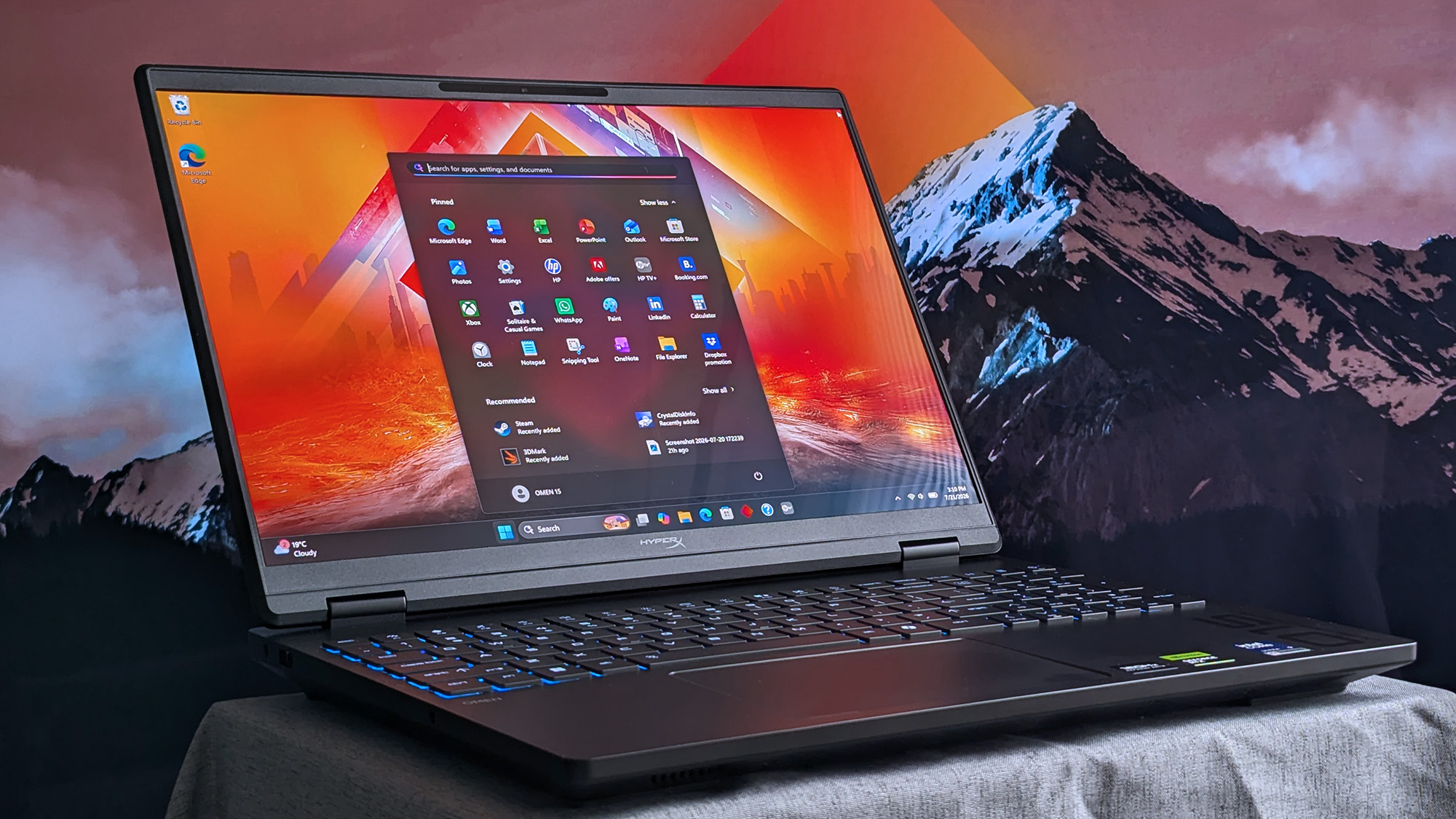 HyperX OMEN 15 laptop with blue&amp;#8209;lit keyboard displaying the Windows 11 start menu over a vivid orange&amp;#8209;red mountain wallpaper, with app tiles and taskbar icons visible on screen