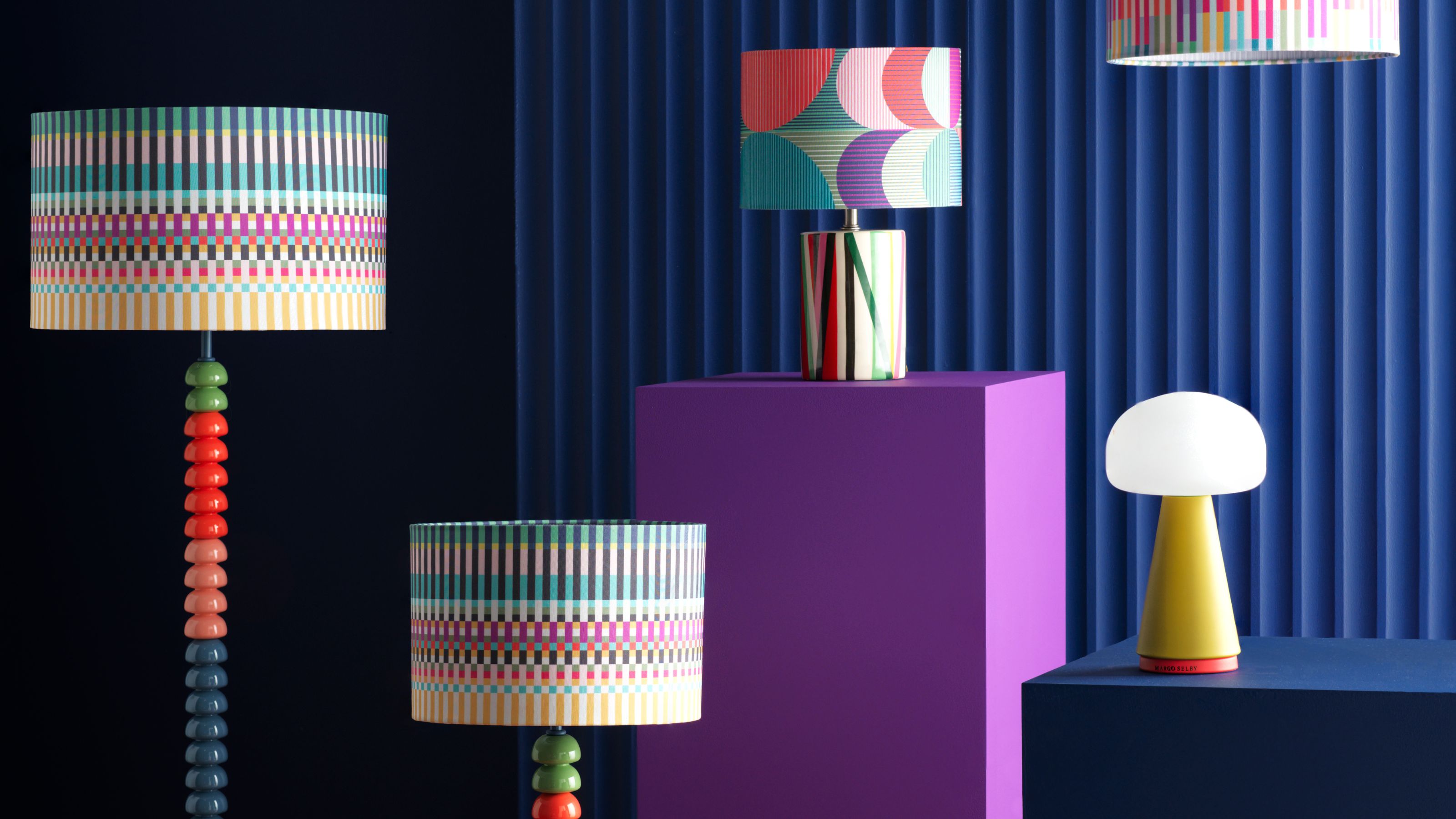 colorful lampshades and bobbin lamp bases on purple pedestal in front of a blue ribbed wall