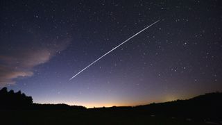 A long white streak is present across the night sky against a background of stars and the horizon below.