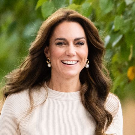 Kate Middleton has long brunette wavy hair and wears a cream sweater and pearl earrings