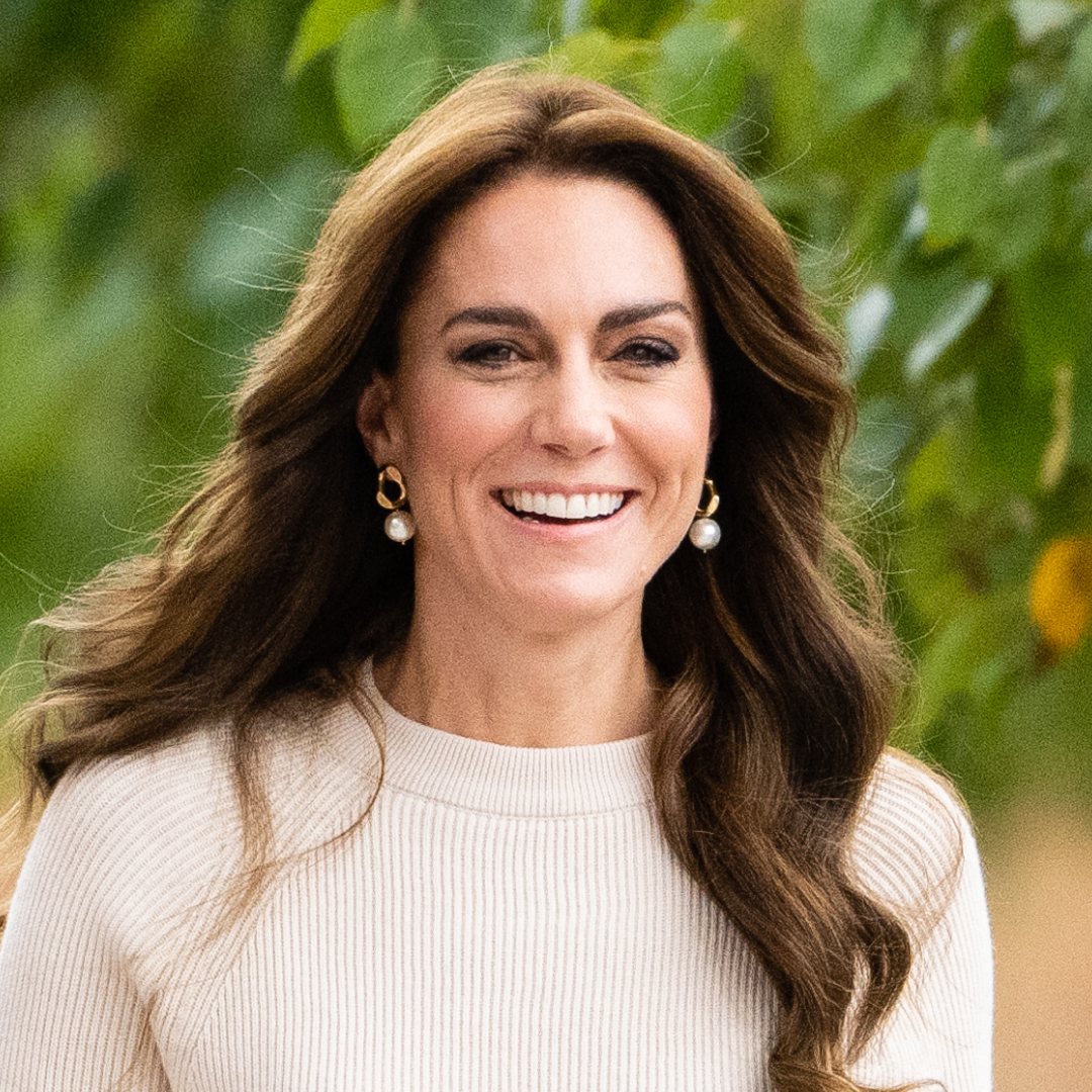 Kate Middleton has long brunette wavy hair and wears a cream sweater and pearl earrings