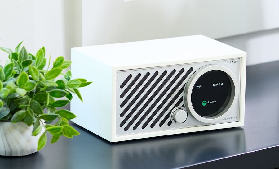 The Tivoli Audio Model One Digital Gen 3 in white on top of a very dark grey/near-black surface with a healthy potted plant beside it.
