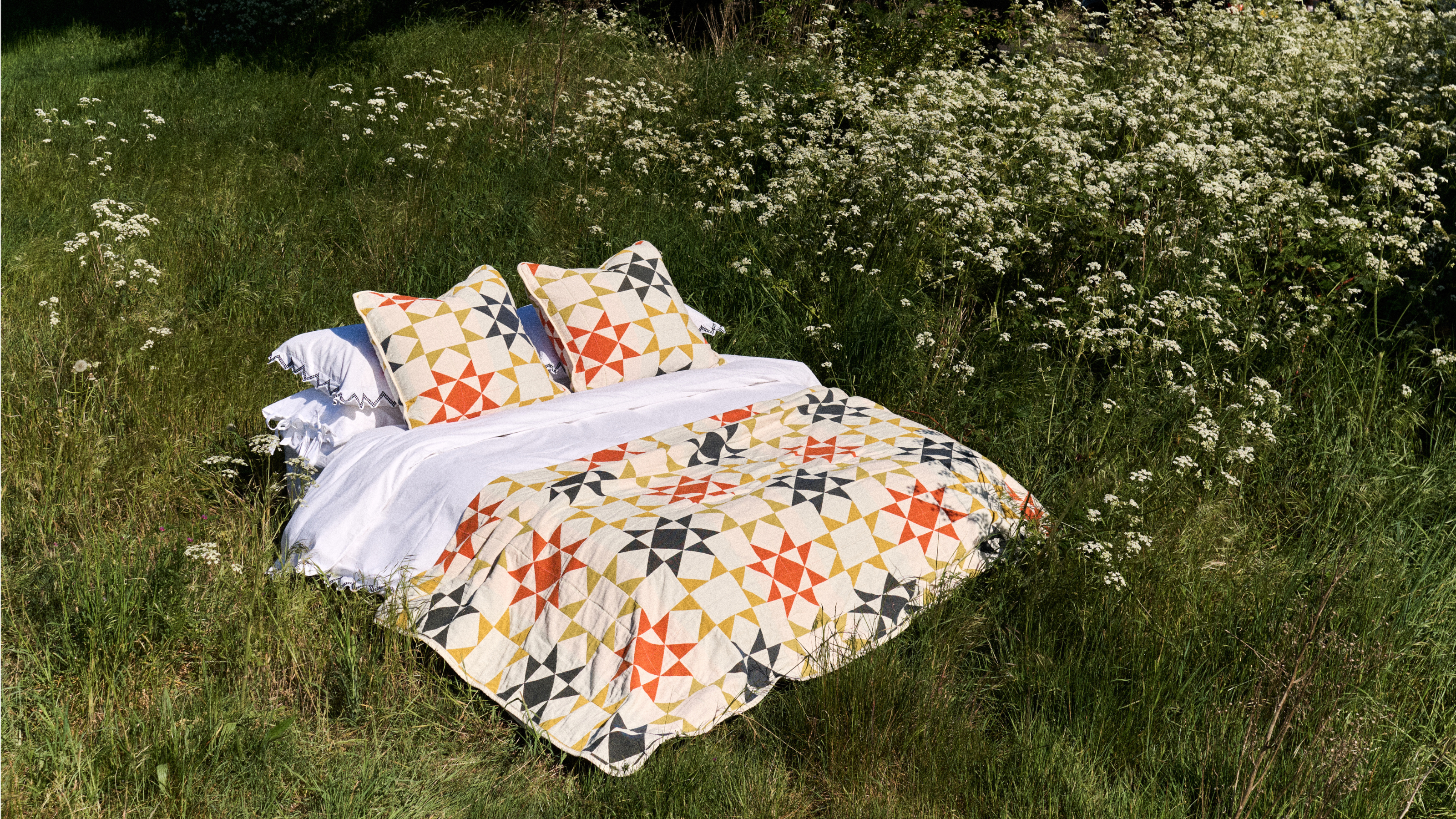 a bed with white sheets and a pinwheel patterned quilt in blue, red, yellow, and off-white resting in a field of tall grass