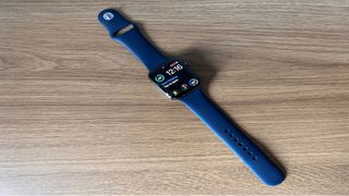 Image shows the Apple Watch Series 7 with a blue strap on a wooden table. The watch face is turned on.