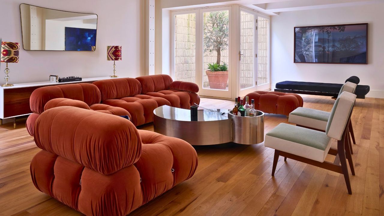 Mid century chairs and cloud burnt orange sofas in minimalist living room, with wooden floors, TV and mirrors on walls, corner terrace with potted tree in view on far left back view