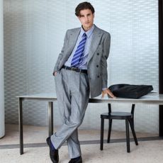 Male model wearing a grey two piece suit with a light blue shirt and darker blue striped tie leaning on a desk for Mytheresa