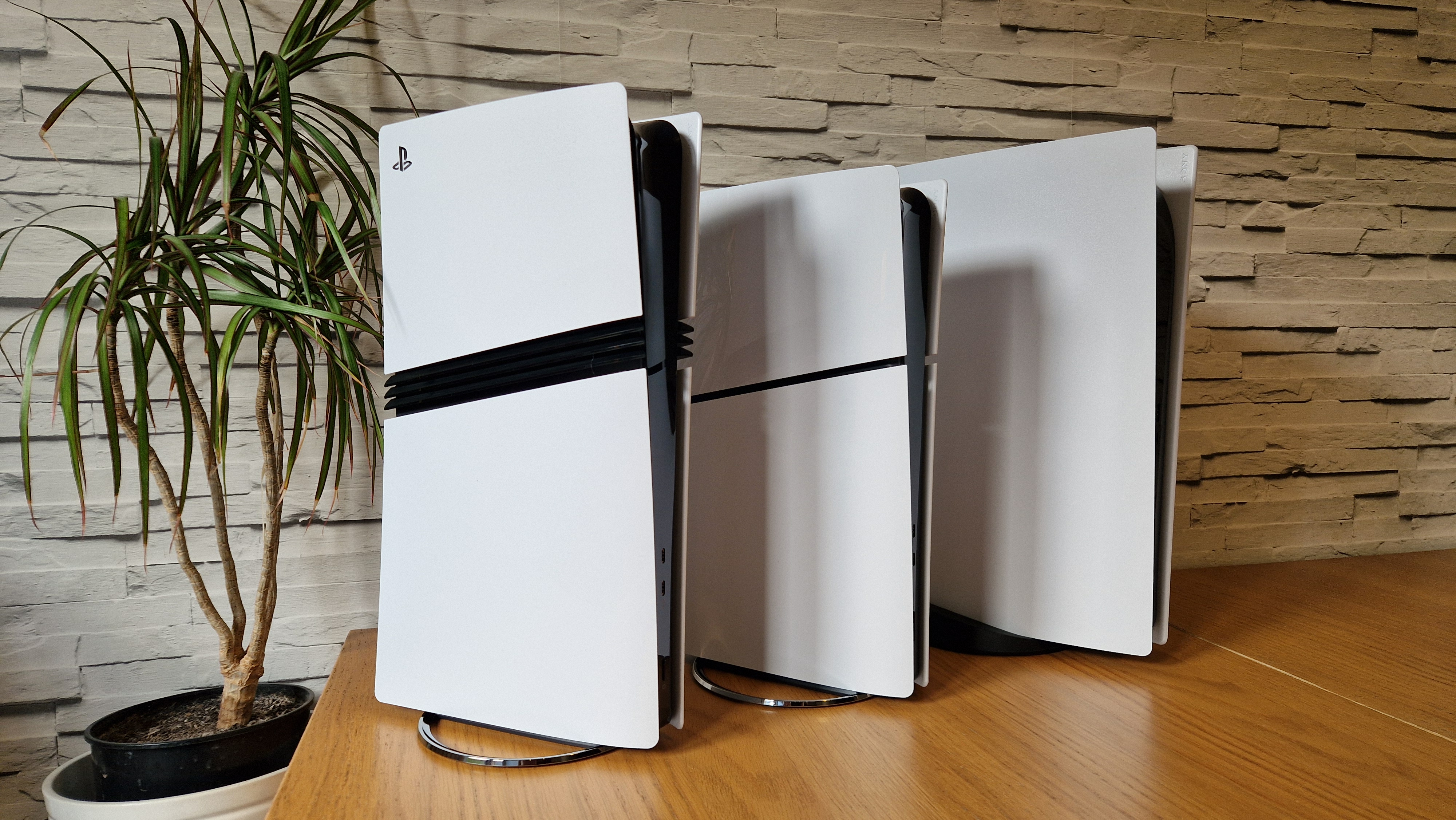 The PS5 Pro console next to a PS5 Slim and launch PS5 on a wooden desk in front of a white brick background and next to a potted plant