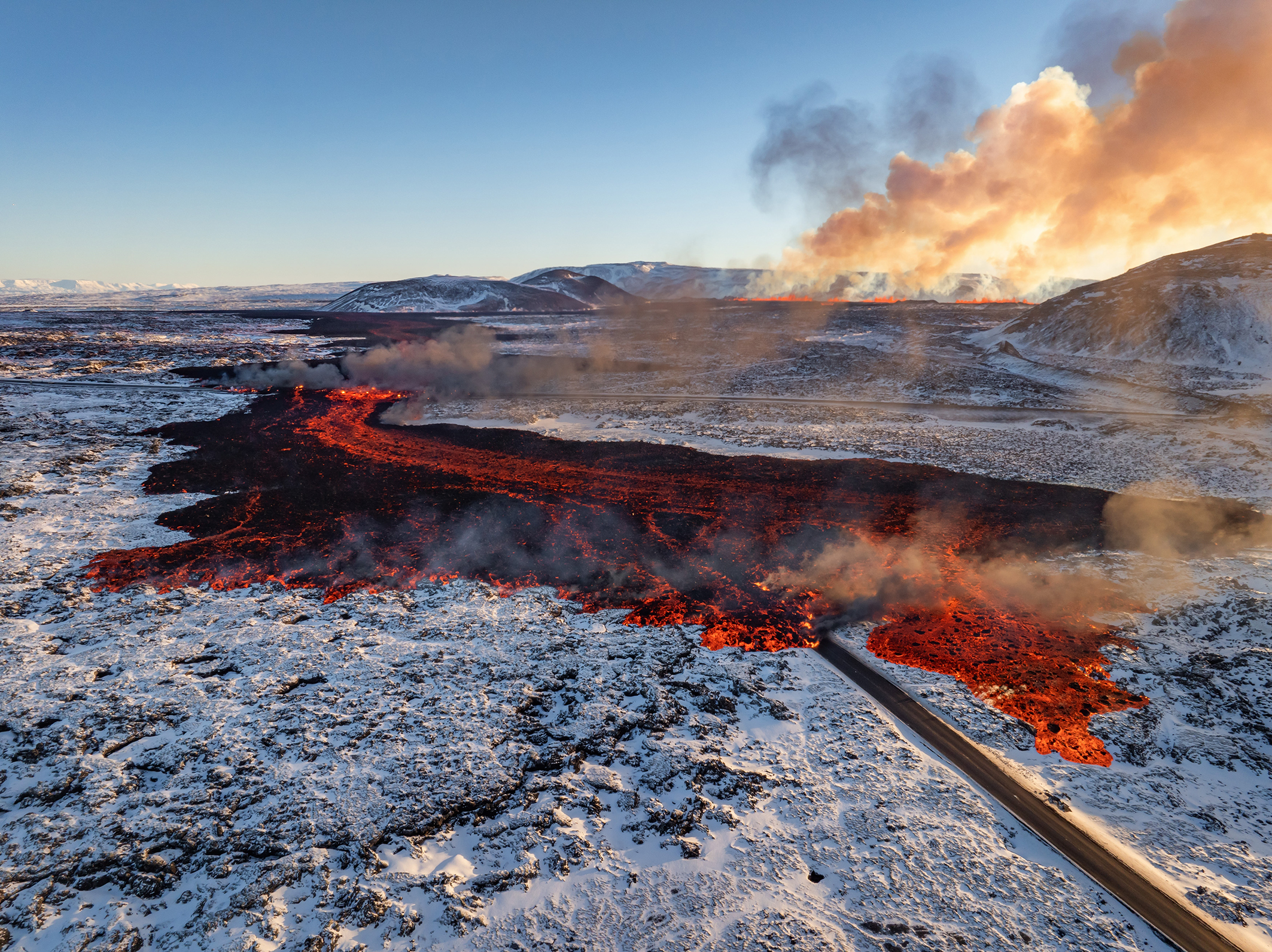 Lava flows across a snowy landscape, with smoke rising in the background and a road visible in the foreground