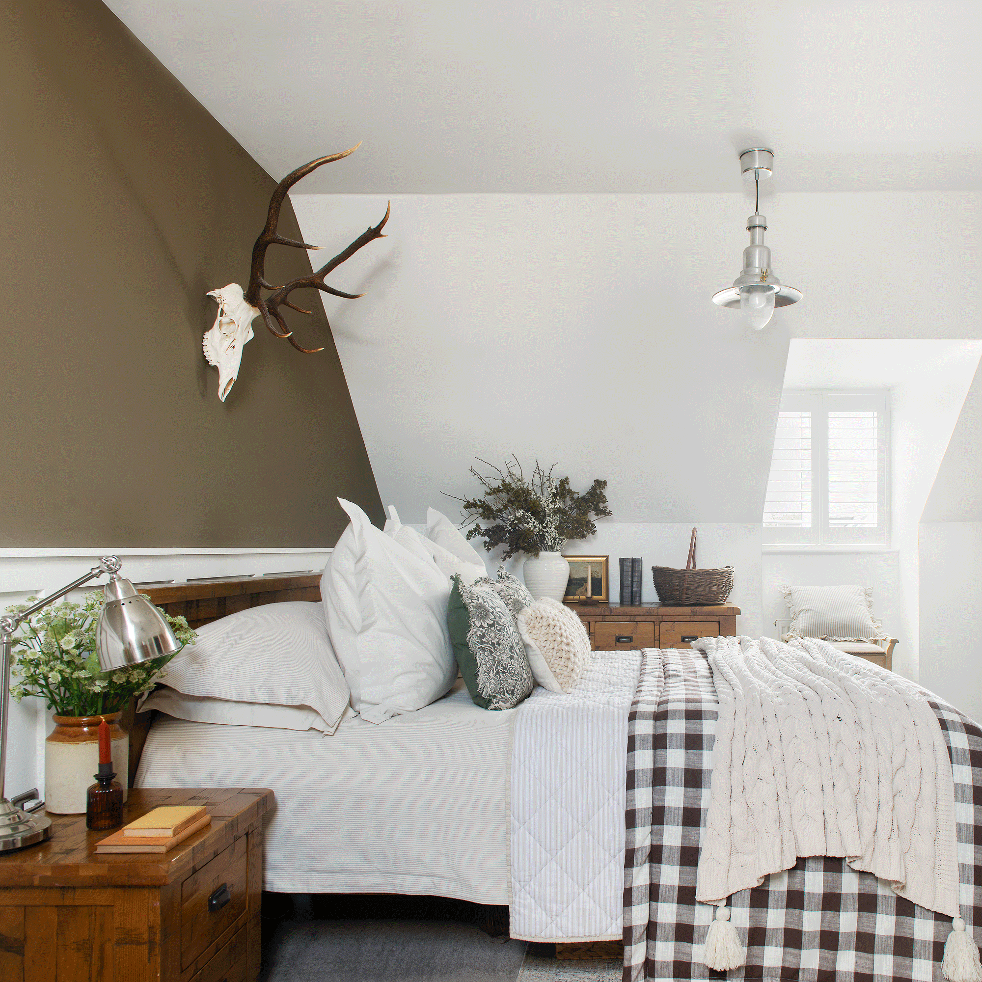 a neutral bedroom with a dormer window and a stag head skull on the wall and check print bed linen on the bed
