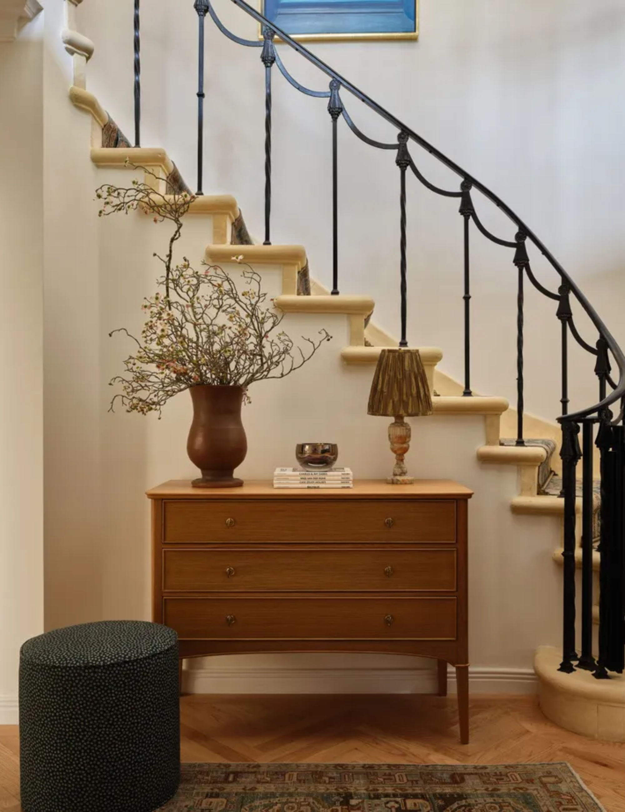 Entryway featuring a curved staircase with a black wrought-iron railing, a mid-century wooden three-drawer dresser, a decorative lamp, and a tall vase with dried branches