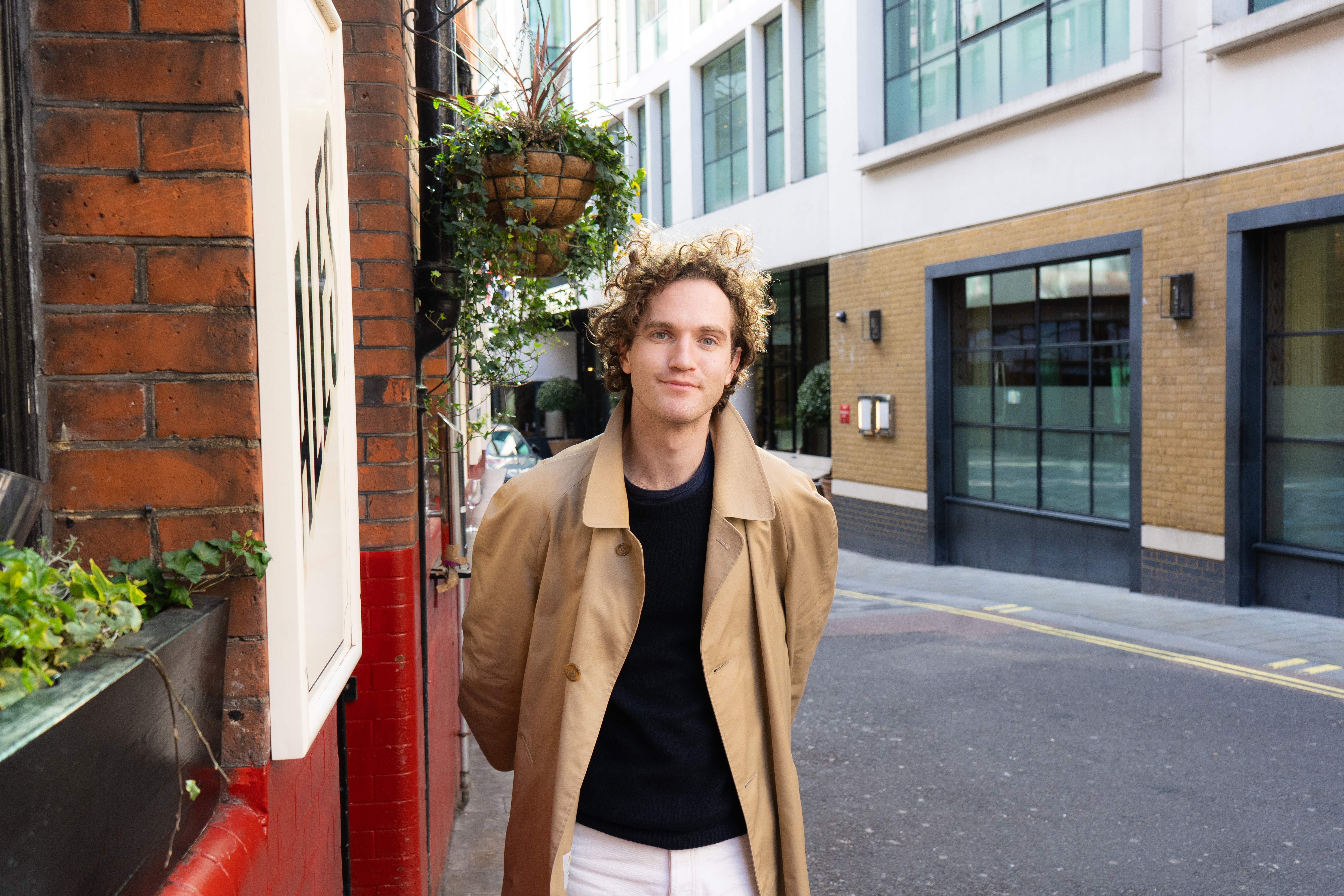 Louis Elton wearing a trench coat, black tee and white trousers out on a sunny day