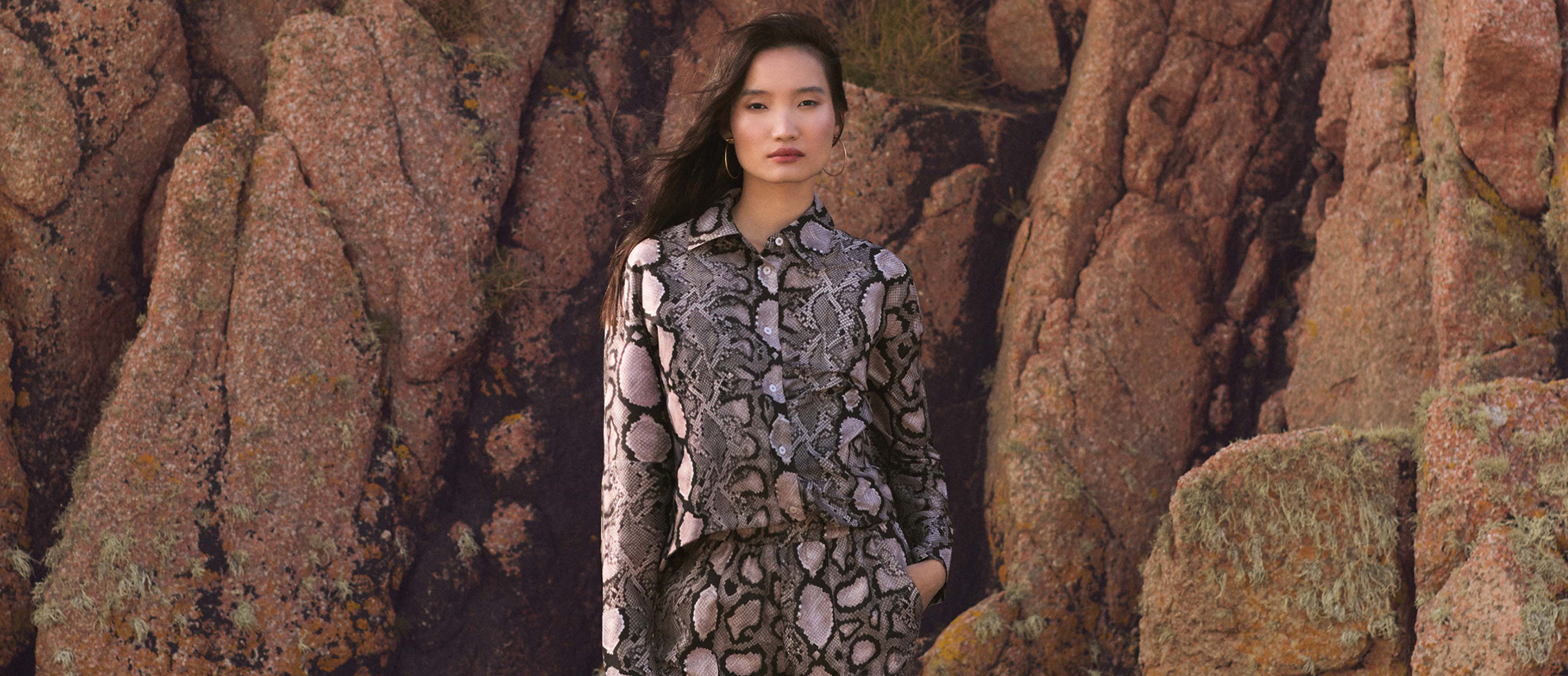 Model wearing an AllSaints snake skin shirt and trouser set stood infront of a rock face 