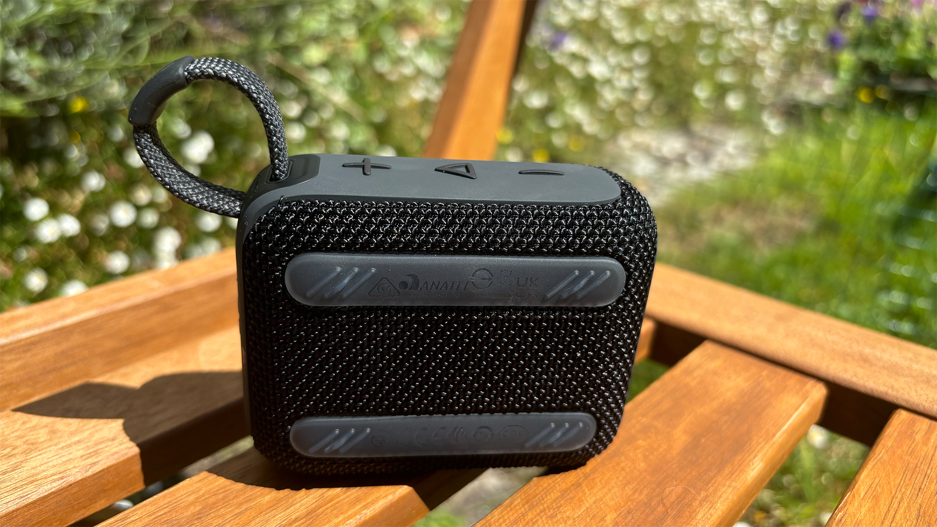 The back of a black JBL Go 4 Bluetooth speaker. The buttons are also visible on top. It is positioned on a wooden garden chair.
