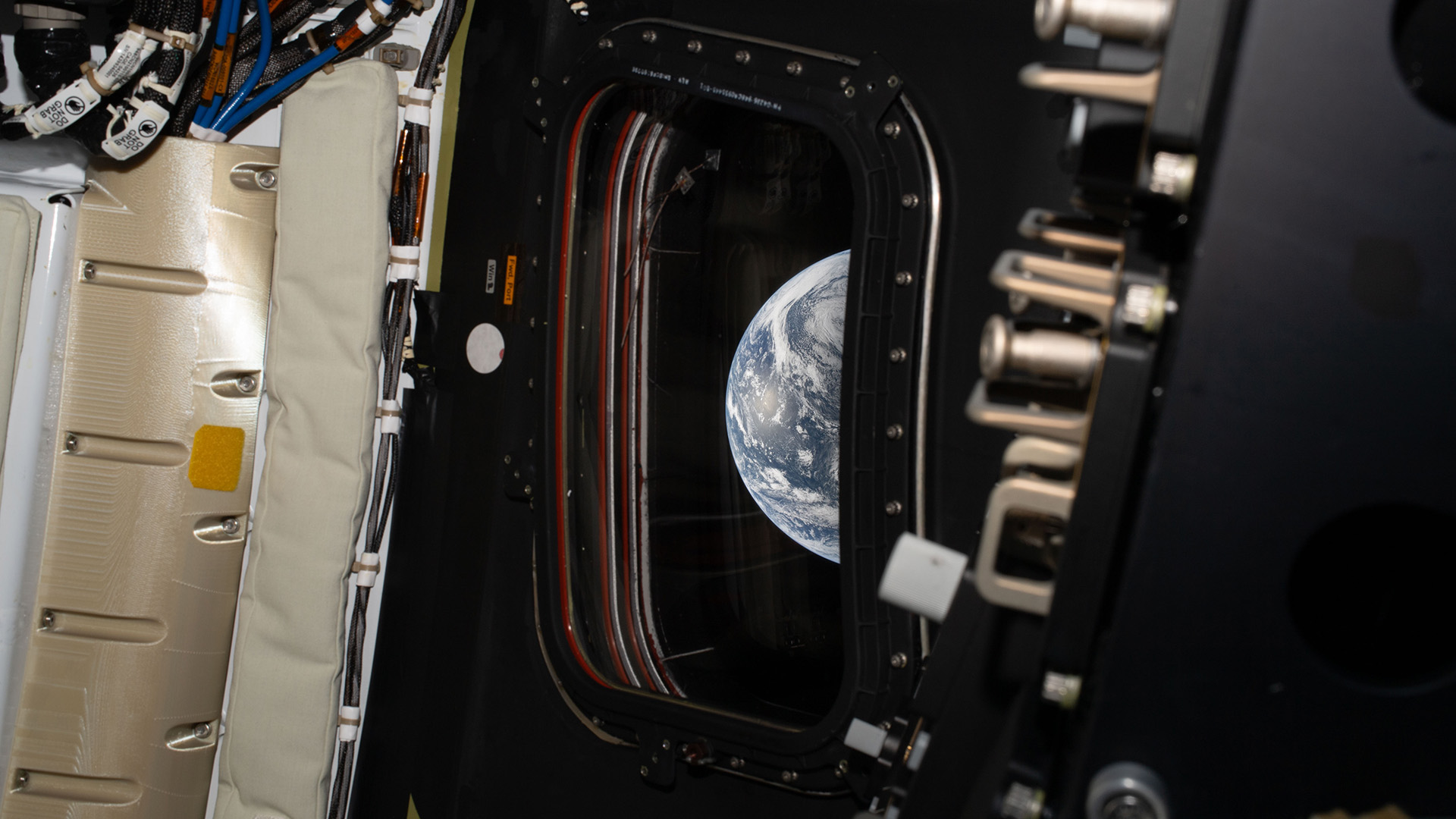 A view of Earth, its blue and white colors, are seen through a rectangular view port window inside a space capsule.
