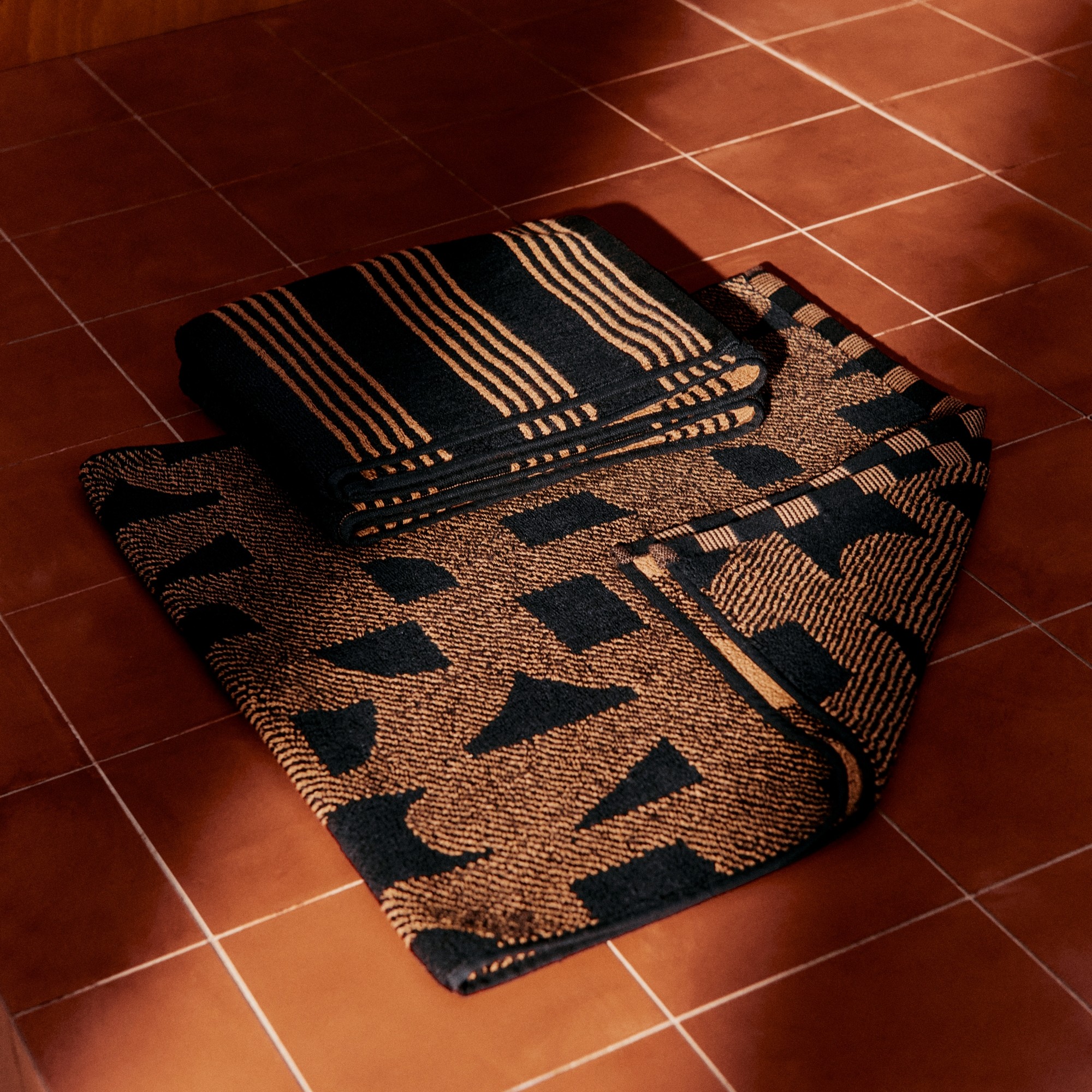 The geometric-pattern towel and striped bath mat from John Cooper's collection for John Lewis laid out on a terracotta tiled floor