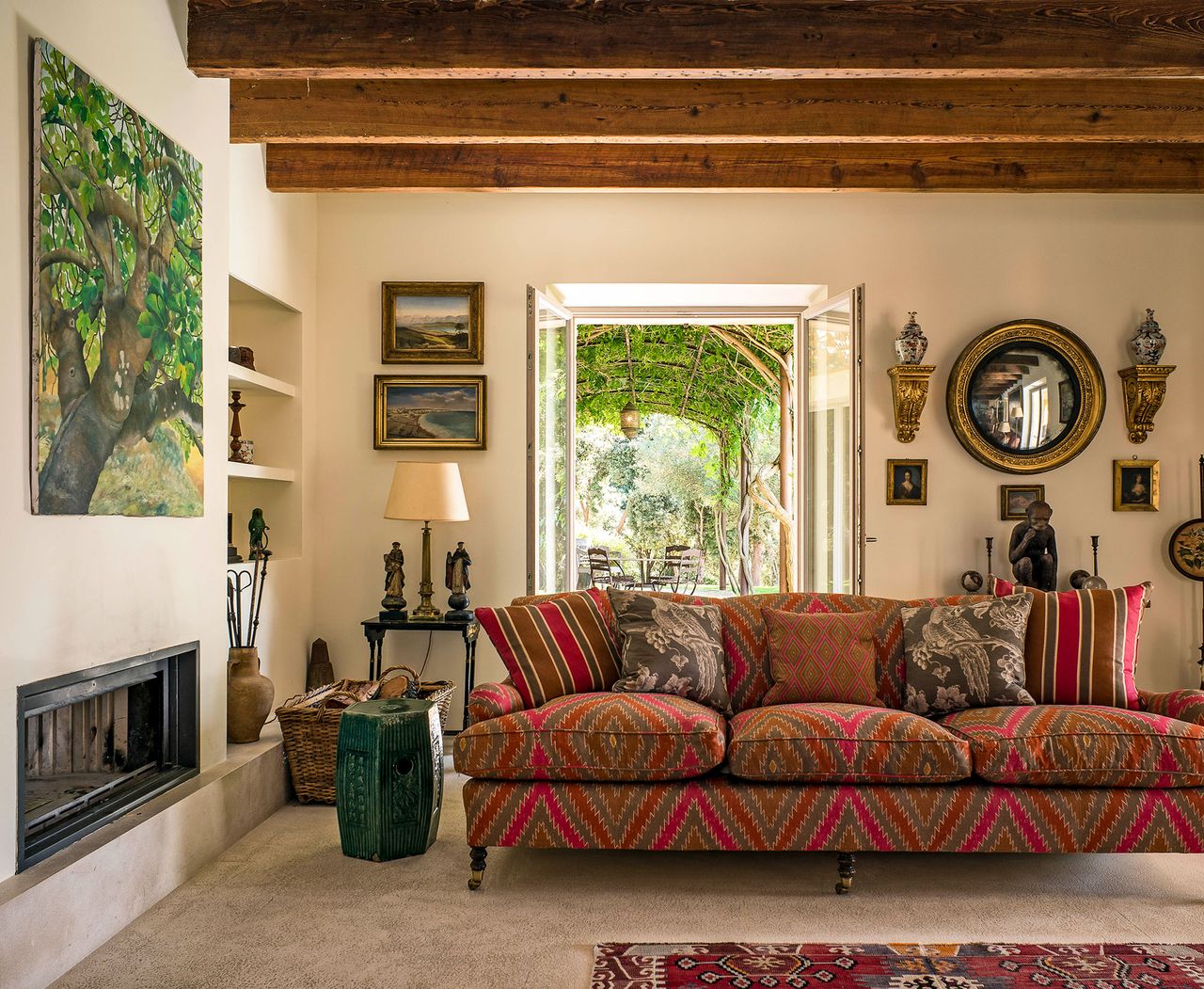 living room with sofa in brick colored diamond print fabric and modern fireplace with large tree painting above and doors to garden arbor open behind an