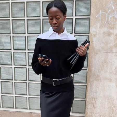 Paris-based fashion influencer Sylvie Mus poses on the sidewalk in a black off-the-shoulder top layered over a button-down shirt along with a belt and satin skirt while looking at her cell phone