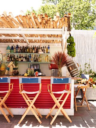 Gilligan's, New York, USA. An outdoor bar with a red counter, blue directors chairs, liquor shelves and a roof made of pieces of wood.