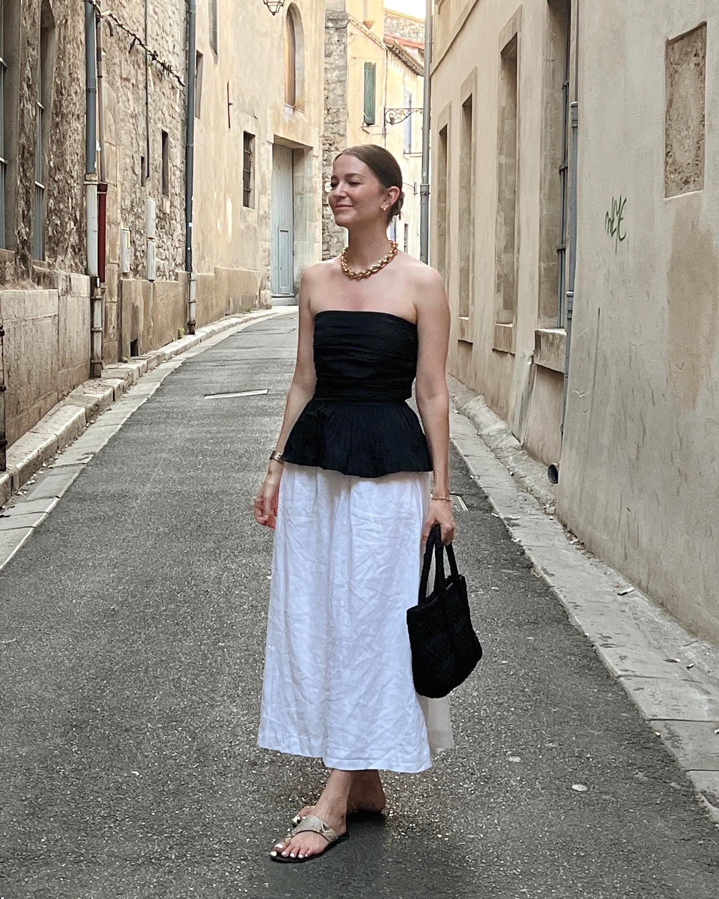 style influencer Marissa Cox poses on a charming street in Arles, France wearing a statement chunky gold chain necklace, a strapless black peplum top, knit black tote bag, white full cotton skirt, and flat snakeskin toe-ring sandals