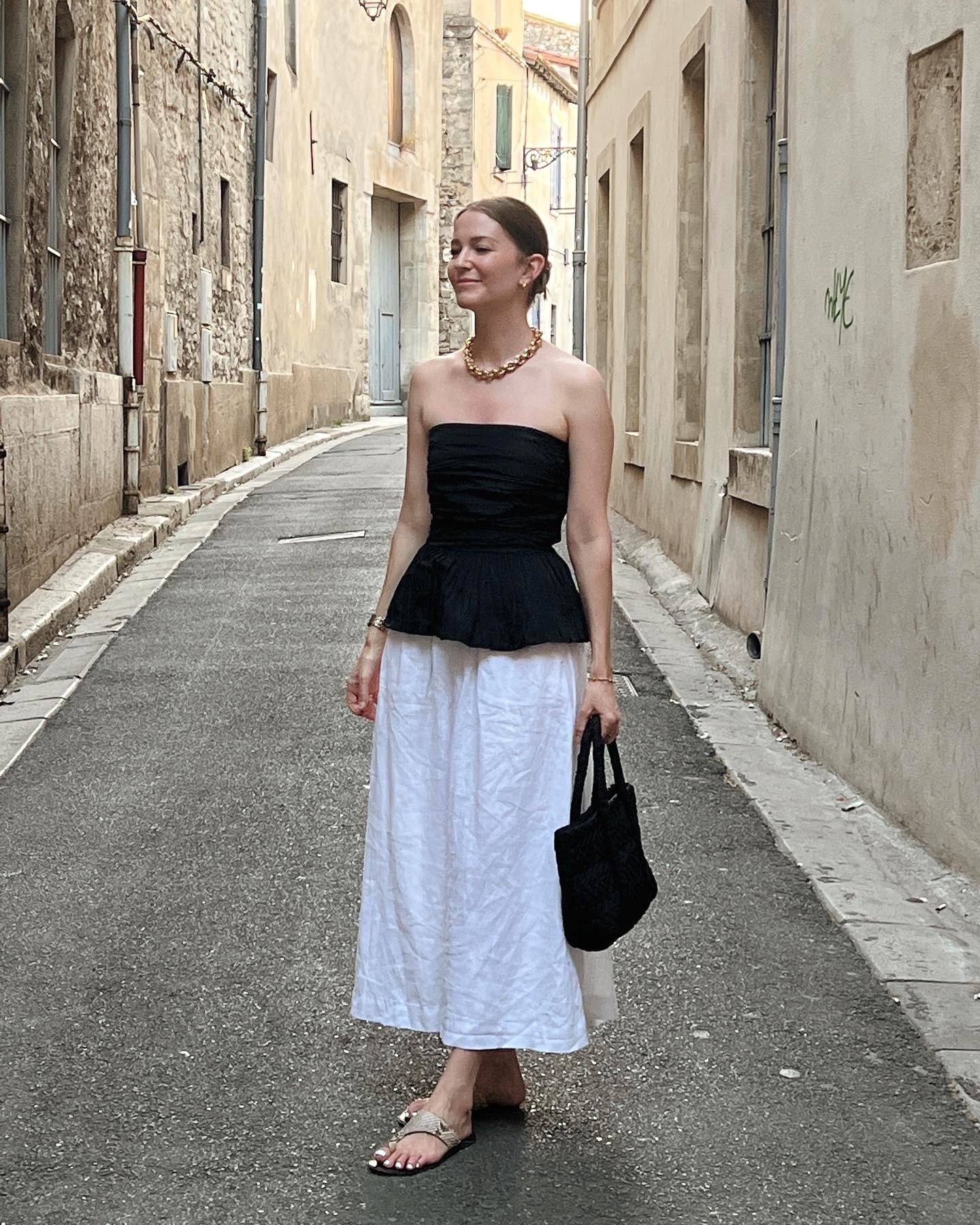 style influencer Marissa Cox poses on a charming street in Arles, France wearing a statement chunky gold chain necklace, a strapless black peplum top, knit black tote bag, white full cotton skirt, and flat snakeskin toe-ring sandals