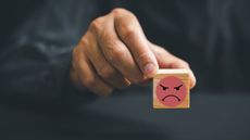 A man's hand holds a wooden block with a scowling emoji on it.