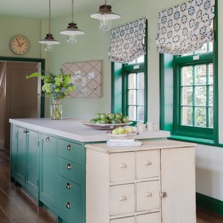 turquoise blue kitchen island and windows, white worktop, vintage unit at end, vintage enamel and glass pendants, green walls, patterned blinds