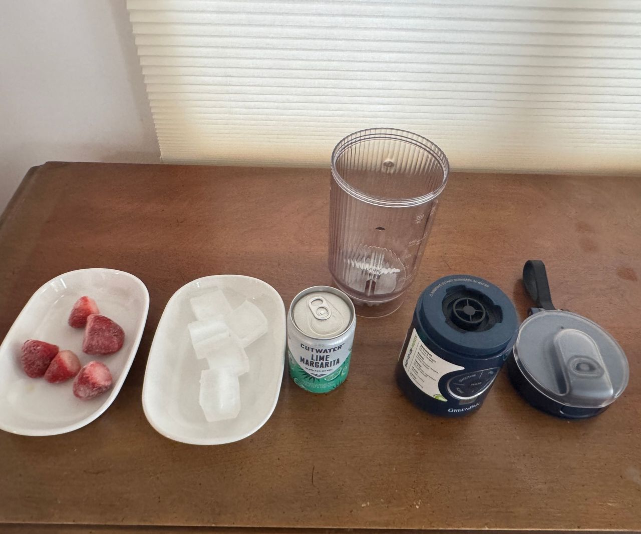 A dismantled GreenPan Spin &amp; Go blender on a wooden table with a plate of frozen strawberries, ice, and a tin of margarita mix.