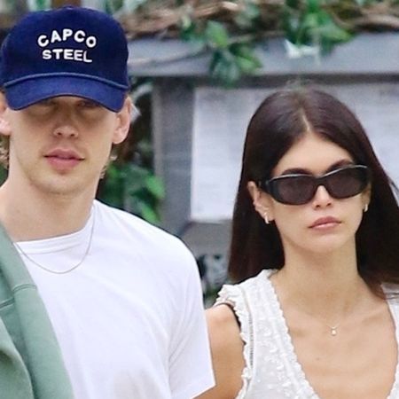 Austin Butler wearing a navy blue hat, white T-shirt, and jeans, with girlfriend Kaia Gerber, wearing a pretty white embroidered top with navy jersey pants, brown ballet flats, and sunglasses walking in NYC.