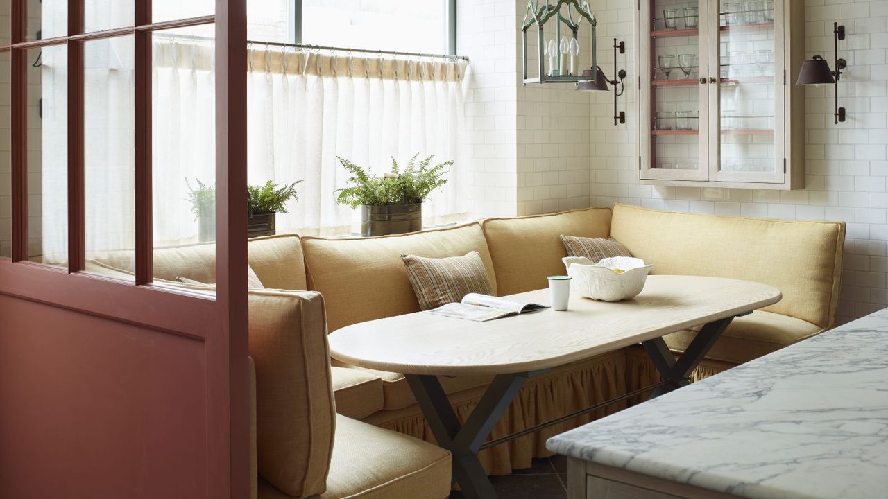 dining nook with a yellow sofa banquette suit with a skirted bottom and a marble table beneath a window with a cafe curtain