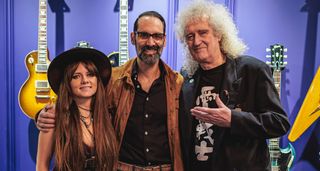 Arielle, Cesar Gueikian and Brian May are pictured at the Gibson Garage London. The wall behind them is painted purple and has Gibson guitars on display.