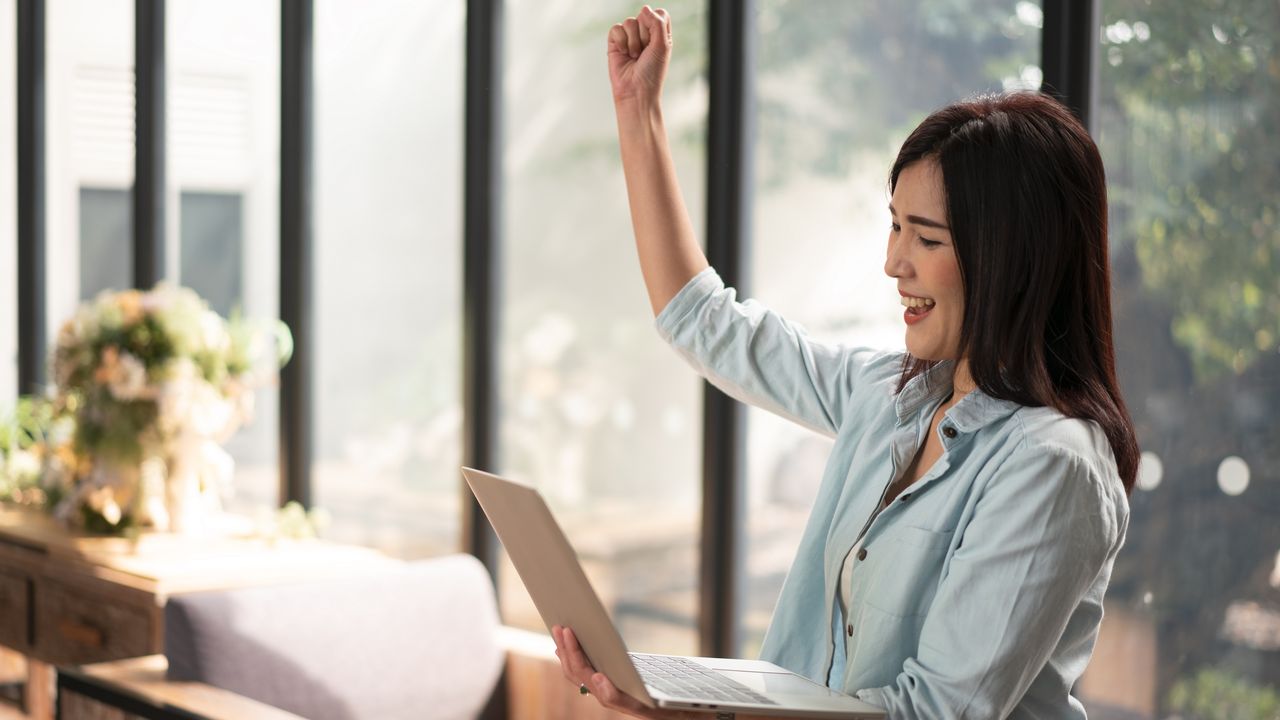 A woman sitting at her desk celebrates her raise while looking at her phone.