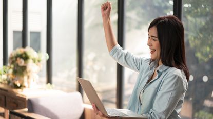 A woman sitting at her desk celebrates her raise while looking at her phone.