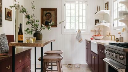 A cottage kitchen with white paneled walls, burgundy cabinets, wooden countertops, and a cozy eat-in area