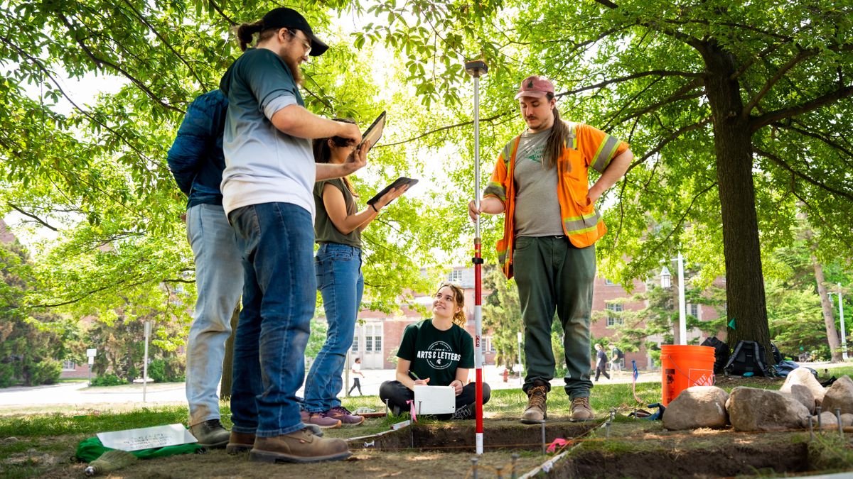 A person holds a long rod in one of two square holes dug into the grass. Three others take notes on paper or iPads.