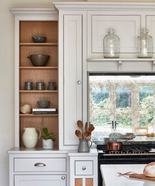 A white kitchen cabinet set surrounding a window, with the cabinet to the left having open shelving with 4 shelves of dark cookware and jars, two glass pendant lights on the ceiling, and a black oven below the window